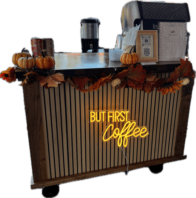 The Azalea Caffeine Collective espresso cart: a wood-slat bar on wheels with a glowing But First Coffee neon sign, dressed in autumn leaves and pumpkins, with the espresso machine and menu on top.