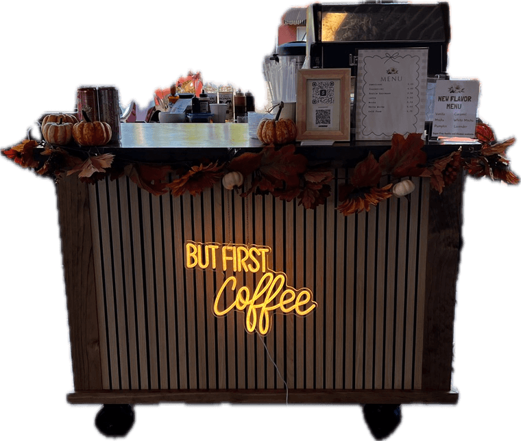 The espresso cart set up for a market day, with its But First Coffee neon sign lit and a fall garland along the counter.