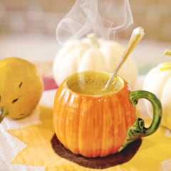 A steaming drink in a pumpkin-shaped mug beside white pumpkins
